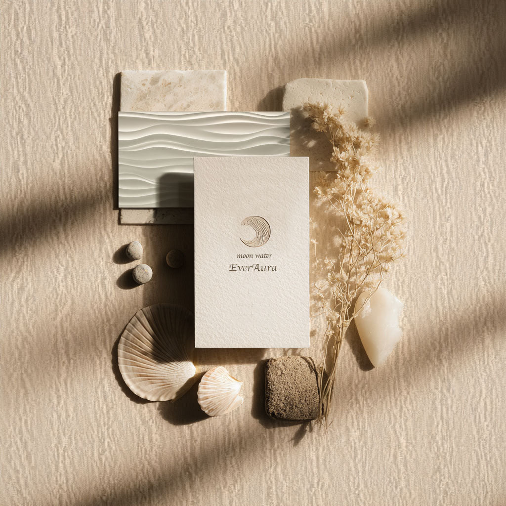 Minimal flat lay of the Moon Water ritual card with a crescent emblem, arranged with seashells, stones, dried flowers, and textured tiles in warm, slanting sunlight.