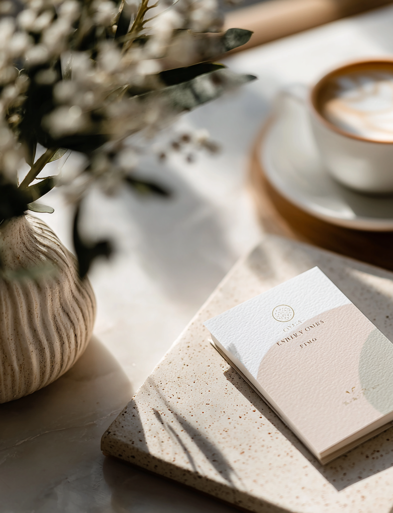 Sunlit tabletop scene with a minimalist journal on a speckled tray beside a cup of latte, with soft-focus flowers and gentle shadows creating a calm morning ritual mood.