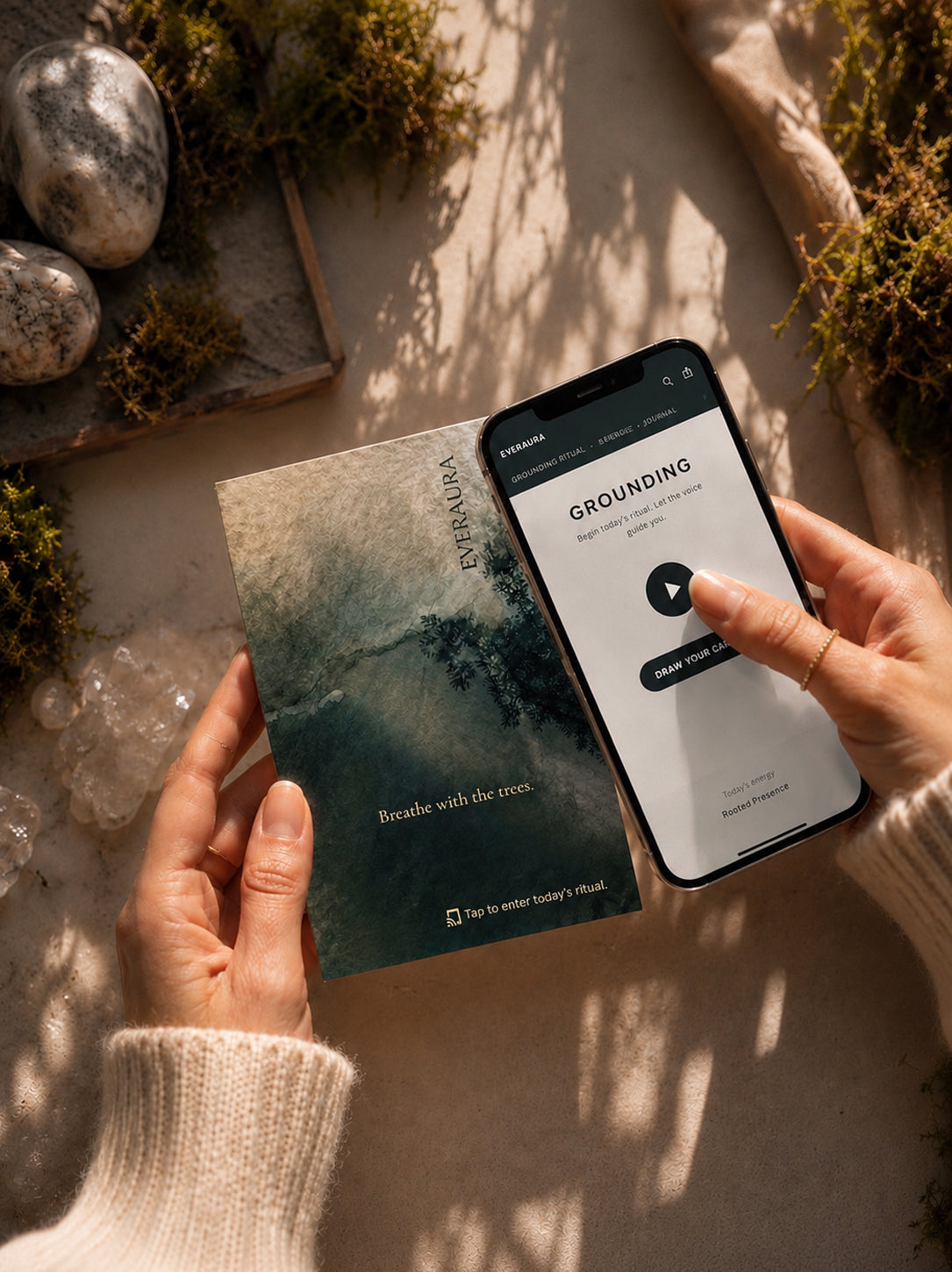 Hands tapping an EverAura Grounding ritual card from the Four Energies Set beside a phone showing the Grounding Daily Ritual. Tap to open a one minute nature meditation.