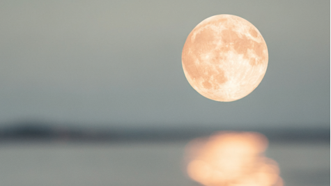 A bright full moon hangs over calm water, with a soft, blurred reflection glowing on the surface below.