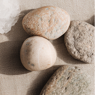 Four smooth stones rest on textured beige fabric in warm light, with a white crystal partially visible in the corner.