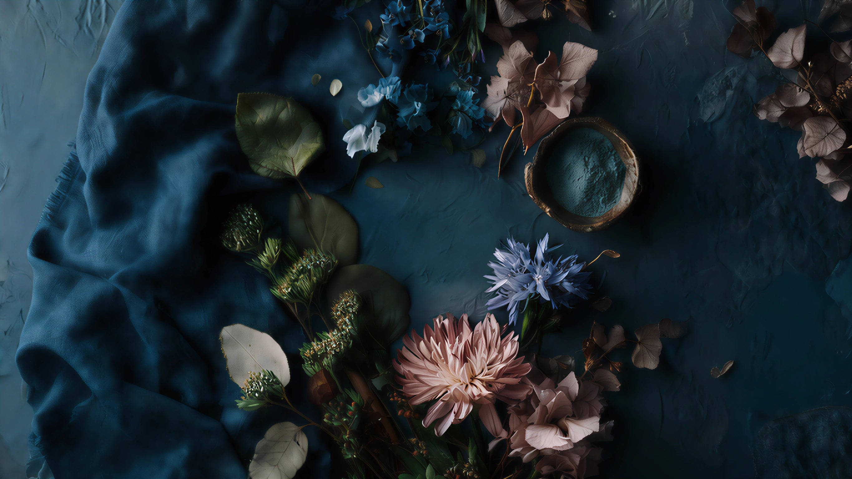 Moody flat lay of blue fabric, dried botanicals, and scattered petals surrounding a small bowl of blue clay powder on a textured dark surface.