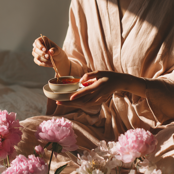 Person in a soft robe holds a small cup and stirs a warm drink with a spoon, surrounded by pink peonies in golden morning light.